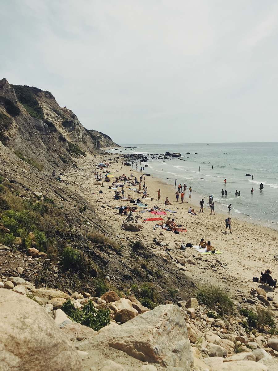 beach people stand on seashore near gray rock formation ocean